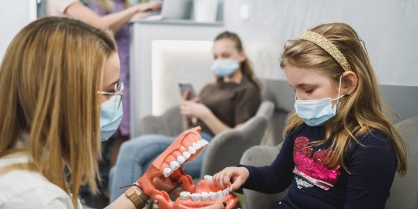 Female dentist and little girl wearing protective face masks while communicating in waiting room.