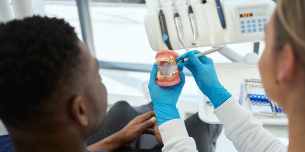 Dentist using sickle probe to show to male client his problems teeth on jaw model, doctor and patient discussing prosthetics, aesthetic stomatology