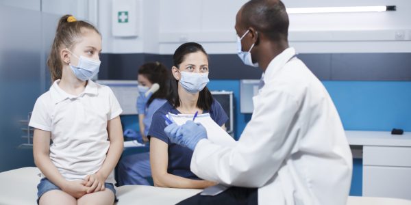 Caucasian mother and her daughter engage in consultation with a healthcare professional. Arican American doctor conducts a medical examination of a young patient, all wearing face masks.